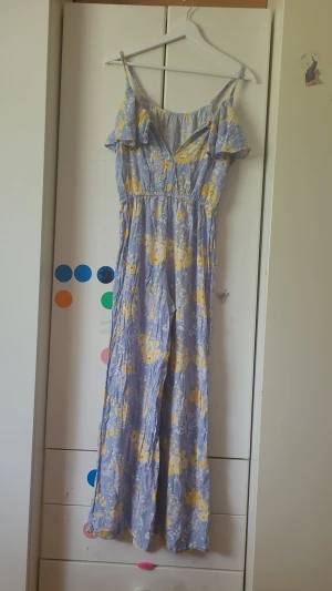 Ljusblå blommig jumpsuit med volang - Supersöt ljusblå jumpsuit med gula och vita blommor. Har tunna axelband, volangdetalj över bysten och resår i midjan för skön passform. Vida ben och mjukt, lätt tyg som känns perfekt för sommaren. Ingen synlig logga eller märke.