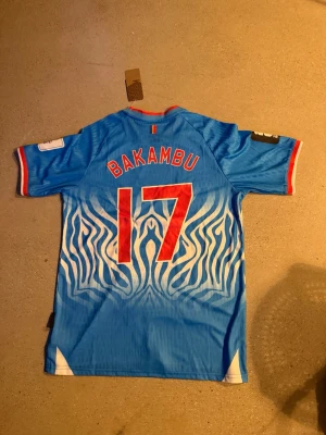 Blå Umbro fotbollströja DR Kongo - Snygg blå fotbollströja från Umbro för DR Kongo med zebramönster i vitt, röda detaljer och nummer 17 samt namnet Bakambu på ryggen. Tröjan har korta ärmar, klubbmärke och flagga på bröstet samt röd kant vid hals och ärmslut.