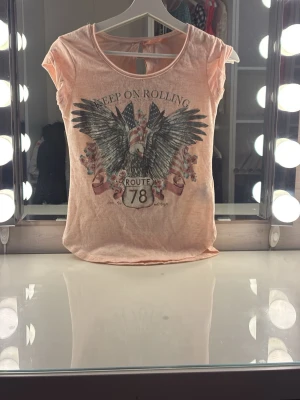 Ljusrosa t-shirt med örntryck - Snygg ljusrosa t-shirt med coolt örntryck, amerikanska flaggor och texten 'Keep on Rolling' samt 'Route 78' framtill. Rund  halsringning och korta ärmar och en mjuk behaglig kvalitet. Detaljer: Under texten "Keep On Rolling" sitter gnistrande strass-stenar som ger trycket ett lyxigt lyft och fångar ljuset fint. T-shirten är i toppskick! 