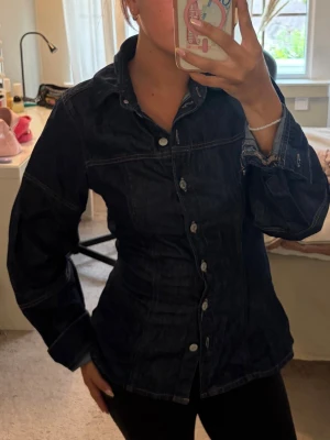 Mörkblå figursydd jeansjacka - Snygg mörkblå jeansjacka med markerad midja och klassisk krage. Jackan har silverfärgade knappar framtill och tydliga sömmar som ger en cool look. Perfekt för dig som vill ha en stilren och trendig jeansjacka i garderoben.