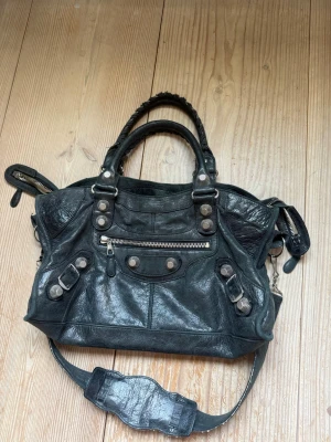 Svart Balenciaga City bag - Cool svart Balenciaga City handväska i mjukt läder med silverfärgade nitar och dragkedjor. Väskan har både handtag och avtagbar axelrem, samt flera fack och detaljer som ger en edgy vibe. Köpt på vestiare collective, helt äkta och checkades hos deras specialister. Den är i använt skick men går att snygga upp hos väskmakaren på NK!