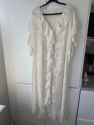 Vit långklänning med volang - Elegant vit långklänning med v-ringning och volangdetaljer längs halsringningen och framtill. Klänningen har ett subtilt mönster i tyget och är tillverkad i ett lätt, mjukt material som ger ett luftigt intryck. Perfekt för sommarens alla tillfällen. Finns en lätt fläck, kan gå bort med fläckborttagning. 