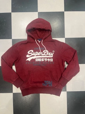 Vinröd hoodie från Superdry - Snygg vinröd hoodie från Superdry med stor vit logga och japansk text på bröstet. Klassisk känguruficka framtill, ribbade muddar och huva med vita dragsnören. Skick enligt bildernaPerfekt för en avslappnad och trendig stil.