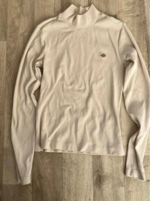 Vit polotopp från Dickies - Vit långärmad polotopp från Dickies med liten logga på bröstet. Topp i mjuk bomull med hög krage och enkel, stilren design. Perfekt att styla lager på lager eller bära som den är.