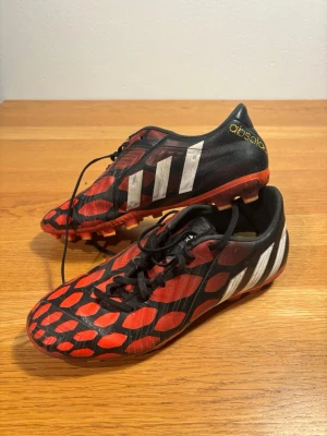 Adidas Predator svart/röd fotbollssko - Säljer ett par Adidas Predator fotbollsskor i svart och rött med vita ränder på sidorna. Skorna har en cool design med röda detaljer och mönster, samt snörning för bra passform. Yttermaterialet är syntet och sulan är anpassad för spel på gräs.