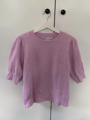Ljuslila stickad topp från ONLY - Söt ljuslila stickad topp från ONLY med korta puffärmar och rund halsringning. Tröjan har ribbade muddar vid ärmslut och nederkant, vilket ger en snygg passform. Perfekt för dig som gillar en feminin och trendig stil.