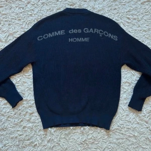 Comme des Garçons- stickad tröja  - Snygg mörkblå stickad tröja från Comme des Garçons HOMME med stor logga i grått på ryggen. Tröjan har rund halsringning, långa ärmar och ribbade muddar. Perfekt för dig som gillar streetwear och exklusiva märken.