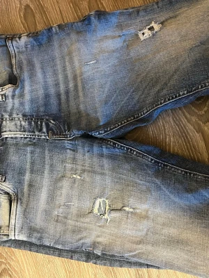Blå slitna jeans med hål - Säljer ett par blå jeans med slitningar och hål på benen. Jeansen har en klassisk femficksmodell och är tillverkade i denim. Perfekta för en avslappnad och trendig look.Skriv för fler frågor och bilder