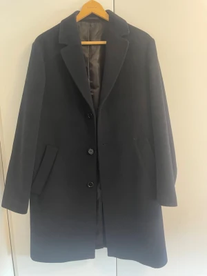 Mörkblå ullkappa med klassisk krage - Stilren mörkblå ullkappa med klassisk krage och tre knappar framtill. Kappan har två snedställda fickor och en rak passform som ger en tidlös look. Perfekt för kyliga dagar och funkar till de flesta outfits. Fodrad insida för extra komfort. Köpt för 1200, använd fåtalet gånger. Det hör ör ett kap!