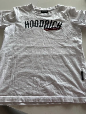 Vit t-shirt från Hoodrich med tryck - Snygg vit t-shirt från Hoodrich med svart tryck över bröstet och en röd detalj. Klassisk passform med rund hals och korta ärmar. Perfekt för en avslappnad streetwear-look. Tillverkad i mjuk bomull för skön känsla hela dagen.