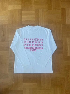 Vit långärmad Maison Margiela t-shirt - Vit långärmad t-shirt från Maison Margiela med ikoniskt tryck i rosa på ryggen. T-shirten har klassisk passform och är tillverkad i mjuk bomull. Perfekt för dig som gillar stilrena plagg med en twist.