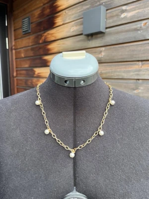Guldfärgat halsband med pärlor - Snyggt halsband med guldfärgad kedja och små vita pärlor som hänger längs länkarna. Kedjan har en klassisk design med rundade länkar och pärlorna ger en elegant touch. Perfekt för att lyfta vilken outfit som helst.