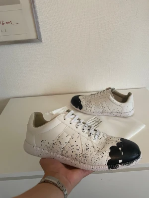 Maison Margiela gats - Säljer ett par extremt snygga och ikoniska Maison Margiela Replica sneakers (German Army Trainers) i den unika ”Paint Drop”-utgåvan. Varje par har ett unikt stänkmönster, vilket gör dem till ett riktigt konstverk på fötterna. De lyfter vilken outfit som helst och är perfekta för dig som vill ha en stilren men samtidigt utmärkande designer-sneaker. Original dustbag (skopåse) medföljer självklart, precis som syns på bilderna! ✨ Detaljer:  Märke: Maison Margiela Paris  Modell: Replica Sneaker / GAT