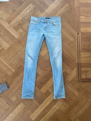 Ljusblå skinny jeans från Nudie - Säljer ett par ljusblå skinny jeans från Nudie Jeans med klassisk femficksdesign och läderpatch bak i midjan. Jeansen har en smal passform och är tillverkade i mjuk denim med lätt tvättad look. Perfekta för en avslappnad och trendig stil.