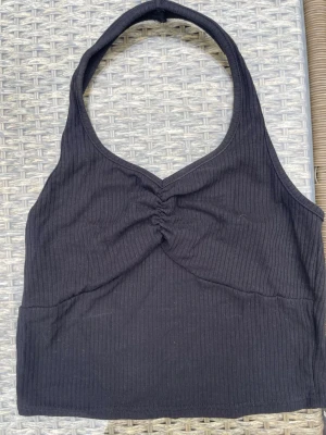 Svart ribbad halterneck topp - Snygg kort  ribbad linnetopp med halterneck och twistad detalj framtill. Perfekt att matcha med  jeans eller kjol för en cool stil.