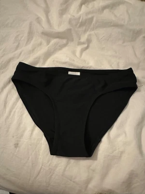 Svarta bikiniunderdelar i enkel modell - Säljer ett par svarta bikiniunderdelar i klassisk, enkel modell. De har en låg midja och är tillverkade i ett stretchigt material som sitter skönt på kroppen. Perfekta för stranddagar eller poolhäng. Storlek 140