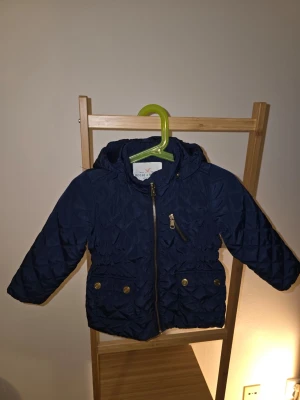 Mörkblå quiltad pufferjacka barn - Snygg mörkblå pufferjacka från lindexl med quiltat mönster, huva och dragkedja framtill. Jackan har två fickor med guldfärgade knappar och en bröstficka med dragkedja.