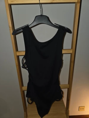 Svart body med snörning i sidan - Säljer en svart body med snygg snörning på sidorna och djup rygg. Bodym har breda axelband och är tillverkad i ett stretchigt material.