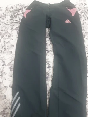 Svarta täckbyxor från Adidas S - Snygga svarta täckbyxor från Adidas med rosa detaljer vid fickorna och klassiska Adidas-loggan på låret. Byxorna har även silvriga ränder på ena benet. Perfekta för kalla dagar och outdoor-aktiviteter.