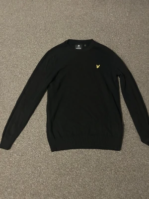 Svart stickad tröja från Lyle & Scott - Klassisk svart stickad tröja från Lyle & Scott med rund halsringning och den ikoniska gula örnloggan broderad på bröstet. Tröjan har långa ärmar och ribbade muddar vid ärmslut och nederkant. Perfekt basplagg för garderoben. Ändå off med den är att den är köpt i storlek M men den sitter som S 