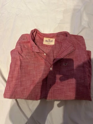 Röd långärmad skjorta från Hollister - Snygg röd skjorta från Hollister med klassisk krage och vita knappar. Skjortan har en liten bröstficka med Hollisters logga broderad. Tillverkad i mjuk bomull och har en avslappnad passform. Perfekt för dig som gillar enkel men stilren design.