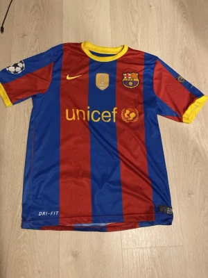 Barcelona Ronaldinho matchtröja - Säljer en klassisk FC Barcelona matchtröja från Nike med Ronaldinho och nummer 10 på ryggen. Tröjan har blå och röda breda ränder, gula detaljer vid krage och ärmslut samt officiella klubbmärken och sponsortryck. Tillverkad i lätt polyester med Dri-FIT-teknologi.