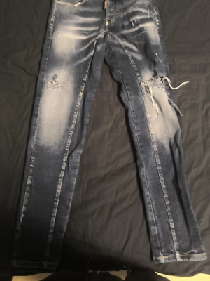 Blå slitna jeans med hål - Säljer ett par blå jeans med slitningar och stora hål på benen för en riktigt trendig och avslappnad look. Jeansen har en tvättad finish med ljusare partier och råa kanter runt hålen. Perfekta för dig som gillar streetstyle och vill sticka ut.