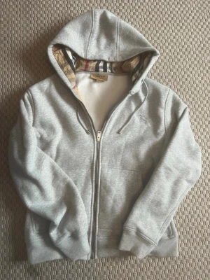 Grå zip hoodie från Burberry - Snygg grå hoodie med dragkedja från Burberry. Klassisk huva med det ikoniska rutmönstret på insidan och broderad logga på bröstet. Tillverkad i mjuk bomullsblandning, ribbade muddar och fickor framtill. Perfekt för en chill och stilren look.