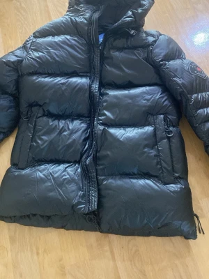 Svart dunjacka från Zavetti Canada - Säljer en svart dunjacka från Zavetti Canada med huva och dragkedja framtill. Jackan har två sidofickor och en stor logga på ärmen. Puffig modell som håller dig varm under kalla dagar. Perfekt för dig som vill ha en stilren vinterjacka.  Köptes för 1500kr men går ej att returnera därför säljer jag den