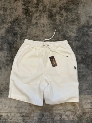 Vita shorts från Polo Ralph Lauren - Snygga vita shorts från Polo Ralph Lauren med elastisk midja och snörning. Klassisk design med fickor på sidorna och en bakficka. Tillverkade i mjuk bomull, perfekta för sommaren. Liten broderad logga på vänster ben.