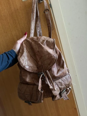 Brun ryggsäck i läder. Vintage stil - Säljer en snygg brun ryggsäck i fejkläder med två framfickor och spännen i metall. Ryggsäcken har justerbara axelband och dragsko under locket för extra säkerhet. Perfekt för dig som vill ha en stilren och praktisk väska till skolan eller stan.