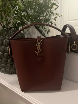 Vinröd bucket bag från Saint Laurent - Säljer en vinröd bucket bag från Saint Laurent i slätt läder med  YSL-spänne i guld framtill.