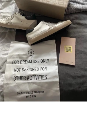 Golden Goose vita sneakers med stjärna - Golden Goose sneakers i vitt läder med grå mockadetaljer och svart häl. Klassisk stjärndesign på sidan och grå skosnören. Snygg och avslappnad look med slitna detaljer. Kommer med originalpåse och box.