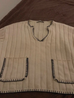 Beige stickad Zara tröja med fickor - Säljer en beige stickad poncho med två stora fickor framtill och svarta dekorativa sömmar längs kanterna. Ponchon har ribbad struktur och en v-ringad hals. Perfekt för lager på lager och chill stil.