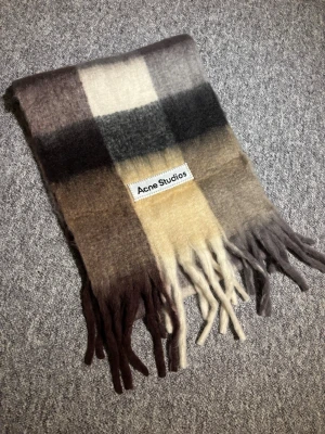 Rutig halsduk från Acne Studios - Snygg rutig halsduk från Acne Studios i mjuk ullblandning. Färgskalan går i beige, brunt, grått och svart med stora blockrutor och långa fransar i ändarna. Perfekt accessoar för att lyfta din höst- eller vinterstil.