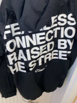 Svart vindjacka med tryck från Faith - Snygg svart vindjacka från Faith med stora vita tryck på ryggen och ärmarna. Texten 'FEARLESS CONNECTION RAISED BY THE STREETS' ger jackan en streetwear-känsla. Jackan har huva, dragkedja och diskret camomönster på axlar och ärmar.