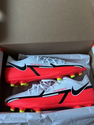 Nike Phantom GT2 fotbollsskor - Snygga Nike Phantom GT2 fotbollsskor i vitt, rött och svart med gula dobbar. Skorna har en modern design med meshmaterial för bra ventilation och greppvänlig yttersula. Perfekta för dig som vill ha kontroll och stil på planen. Aldrig använda.