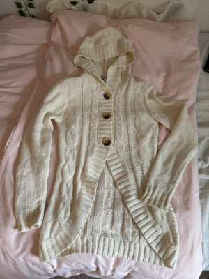Vacker gullig stickad kofta med luva och knappar - Vintage mysig vit/creme stickad lång kofta med huva och stora träknappar framtill. Koftan har kabelstickat mönster, ribbade muddar och en omlottliknande öppning. Perfekt för lager på lager och chill dagar! Så mysig och passar utmärkt till alla plagg❣️Inget stickande material (sista bilden=material). Barnstorlek men passar XS🩷Aldrig använd av mig, hittade i garderoben! 