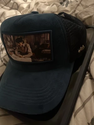 Maggiore keps, (Coco Chanel) - Snygg blå truckerkeps från Maggiore med mesh på sidorna och ett stort tygmärke framtill med konstmotiv. Kepsen har böjd skärm och justerbar baksida. Perfekt för dig som vill sticka ut med en unik accessoar.