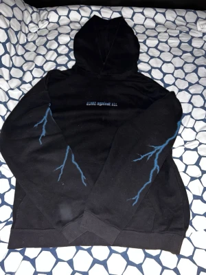 Svart hoodie med blå blixtar & fjäril - Svart hoodie med trycket 'ALONE against ALL' på bröstet och blå blixtar längs ärmarna. Stor ryggdesign med texten 'LIVE OR DIE' och en blå fjäril i eld. Perfekt för dig som gillar streetwear och unika prints.