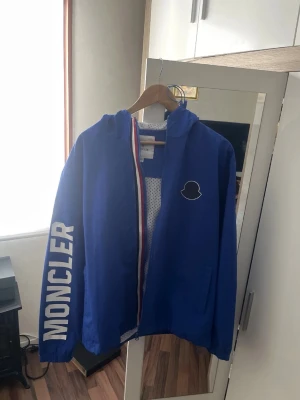 Moncler vindjacka  - Snygg blå vindjacka från Moncler med vit logga på bröstet och stort MONCLER-tryck på ena ärmen. Finns inte många gjorda denna modell vill byta den helst mot en moncler cardigan Strl L men kan sälja den oxå helt ny oanvänd är jackan 