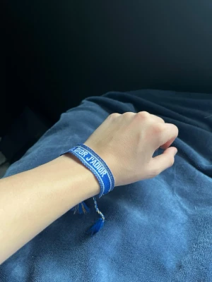 Blått tygarmband Christian Dior - Snyggt blått tygarmband från Christian Dior med texten 'DIOR J'ADIOR' i vitt. Armbandet har dekorativa tofsar i ändarna och justerbar knytning. Perfekt accessoar för att lyfta din outfit med en exklusiv touch.