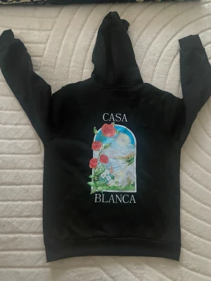 Svart Casa Blanca hoodie med tryck - Säljer en svart hoodie från Casa Blanca med färgglatt tryck av rosor och landskap på både bröstet och ryggen. Hoodien har stor huva och magficka framtill. Perfekt för dig som vill ha en unik och stilren streetwear-look.