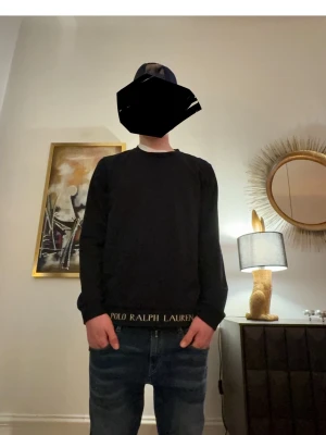 Svart tröja från Polo Ralph Lauren - Säljer en svart långärmad tröja från Polo Ralph Lauren med rund hals och diskret logga i guldtext vid nederkanten. Tröjan har en klassisk passform och är perfekt för en clean och trendig look