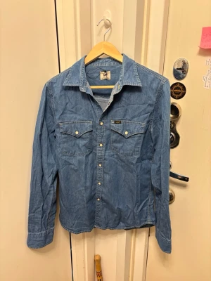 Blå jeansskjorta från Lee - Klassisk blå jeansskjorta från Lee med två bröstfickor och knappar framtill. Skjortan har lång ärm, normal passform och är gjord i bomull. Perfekt för en avslappnad och cool stil. Funkar perfekt för varje dag användning men också för finare situationer! 
