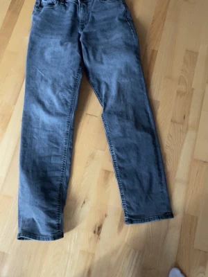 Grå raka jeans med hög midja - Säljer ett par grå jeans med rak passform och hög midja. Klassisk femficksmodell med dragkedja och knapp framtill. Jeansen har en avslappnad look och är tillverkade i denim. Perfekta för dig som gillar en enkel och stilren stil.