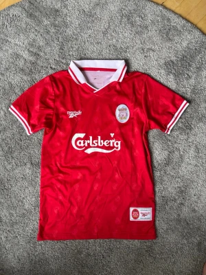 Liverpool retro matchtröja Reebok - Klassisk röd Liverpool FC matchtröja från Reebok med vit krage och detaljer. Carlsberg-logga på bröstet och klubbmärke på vänster sida. Tillverkad i lätt och ventilerande material, perfekt för fotboll eller samling.