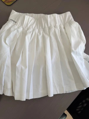 Vit plisserad tenniskjol från Adidas - Snygg vit tenniskjol från Adidas med plisserad design och bred resår i midjan. Kjolen har diskret Adidas-logga framtill och är tillverkad i ett lätt och stretchigt material som passar perfekt för sport som tennis/ padel. Använd fåtal gånger.