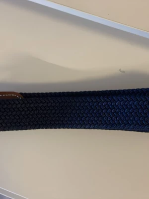 Mörkblått flätat bälte - Snyggt mörkblått flätat bälte med detaljer i brunt läder. Bältet har en elastisk känsla och passar perfekt till jeans eller chinos. Klassisk design som funkar till många olika stilar.