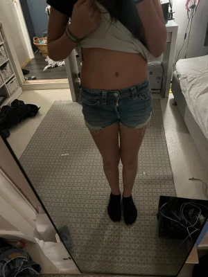 Blå jeansshorts med fransar - Säljer ett par klassiska blå jeansshorts med råa fransar i bensluten och slitna detaljer. Shortsen har normal passform, knappgylf och fickor både fram och bak. Perfekta till sommaren och lätta att matcha med olika toppar.